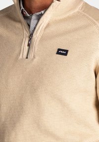 Beige pullover met een texturen breipatroon, voorzien van een kraag met korte rits en een zwart NZA logo patch op de borst.