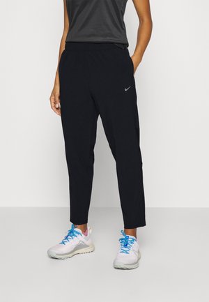 Personne portant un pantalon de sport noir Nike et des chaussures de course blanches avec des lacets bleus, debout devant un fond uni.