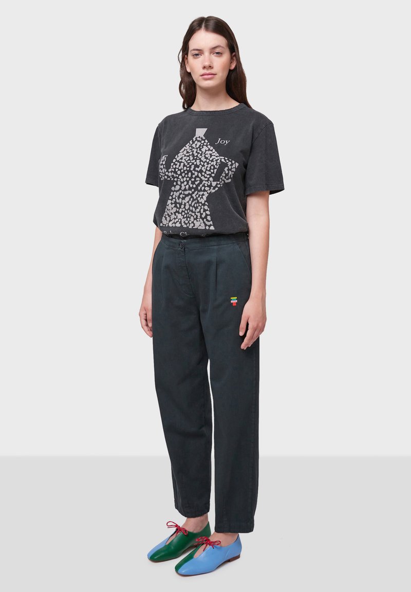 T-shirt grigia con motivo astratto bianco e testo "Joy", abbinata a pantaloni verde scuro con un accento multicolore e scarpe bicolore.