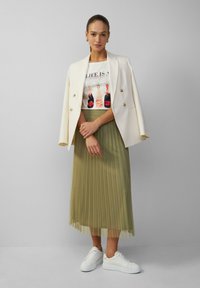 Witte blazer over een wit graphic t-shirt met afbeeldingen van flessen, gecombineerd met een geplooide groene rok en witte sneakers. Soepele texturen en gelaagd ontwerp.