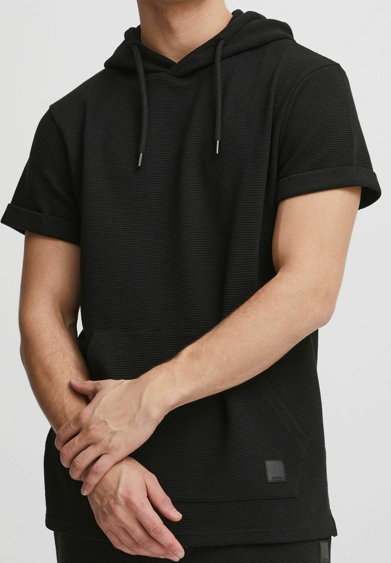 Homme portant un t-shirt à capuche noir à manches courtes retroussées, joignant les mains devant un fond uni.
