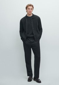 Černý žebrovaný cardigan přes tmavý svetr, v kombinaci s tmavými kalhotami a černými botami. Jednoduchý design s knoflíkovým zapínáním a minimálními akcenty.