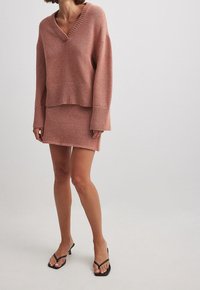 Pull en tricot couleur rose avec un col en V et un ourlet côtelé, associé à une jupe ajustée assortie. Le mannequin porte des sandales à talons noires à brides.