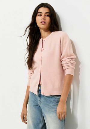 Ung kvinde med langt mørkt hår iført en lyserød cardigan med knapper og lyseblå jeans, der står op ad en hvid væg.