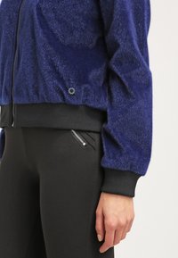Personne portant une veste zippée bleu marine texturée avec des poignets côtelés noirs et un pantalon noir doté d'une petite poche zippée.