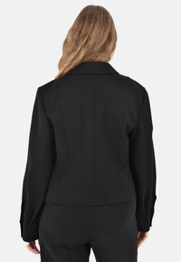 Giacca nera su misura con una silhouette strutturata, ampi risvolti e taglio corto. Presenta dettagli con cuciture e polsini con bottoni.