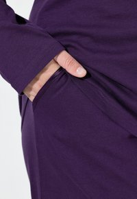 Fialový dlouhý rukáv s hladkým povrchem má boční kapsu, ukazuje přiléhavý design a čistý, jednoduchý střih.