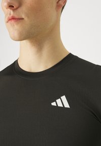 Primer plano de una persona que lleva una camiseta negra con un pequeño logo blanco de Adidas en el pecho, mirando ligeramente hacia la izquierda sobre un fondo liso.