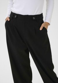 Pantalons noirs sur mesure avec une texture lisse, deux plis avant, des poches latérales et des accents de boutons noirs sur la taille.