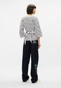 Ριγέ, ελαφρύ λευκό και ναυτικό μπλουζάκι με δεμένο πίσω μέρος; συνδυασμένο με σκούρα τζιν παντελόνια που διαθέτουν λουλουδάτη κεντητική στο αριστερό πόδι.