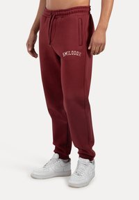Bordeauxkleurige joggers met elastische tailleband, voorzien van zijzakken en witte "SMILODOX" tekst. Gecombineerd met witte sneakers.