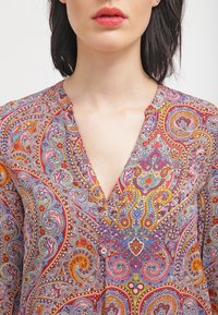 Barevná blůza s paisley vzorem, která má výstřih do V a knoflíky, s živými odstíny červené a modré a složitými detaily po celém materiálu.