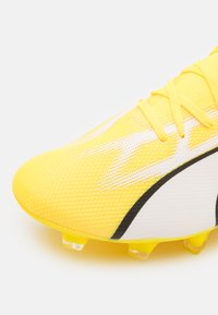 Botín de fútbol amarillo brillante con parte superior de malla texturada, acentos blancos y un diseño de líneas negras. Presenta tacos transparentes para mayor agarre.