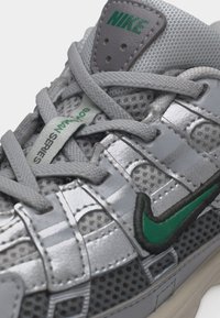 Gros plan sur une basket Nike grise et argentée, avec un logo vert, un matériau en maille et une étiquette "Bowerman Series" sur la languette sous les lacets gris.