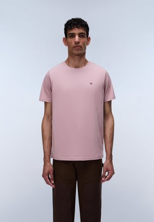 Mann mit kurzen dunklen Haaren, der ein schlichtes pinkes T-Shirt und dunkelbraune Hosen trägt, steht vor einem hellgrauen Hintergrund.