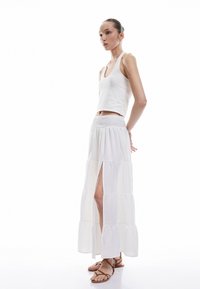 Falda maxi blanca con volantes y cintura fruncida, con aberturas laterales, combinada con una camiseta ajustada blanca y sandalias planas marrones. Textura suave.