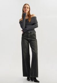 Dunkelgrauer, schulterfreier Rippstrickpullover kombiniert mit hochtaillierten, weit geschnittenen schwarzen Denim-Jeans und schwarzen Stiefeletten mit Absatz.