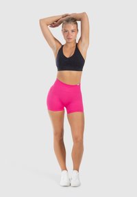 Zwarte sportbeha met een V-hals en ruching-detalje, gecombineerd met hooggetailleerde felroze shorts. Model draagt witte sportschoenen.