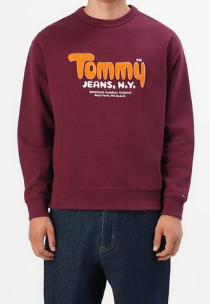 Osoba w bordowej bluzie z nadrukiem „Tommy Jeans, N.Y.” w kolorze pomarańczowym i białym oraz w niebieskich dżinsach, stojąca na jednolitym białym tle.