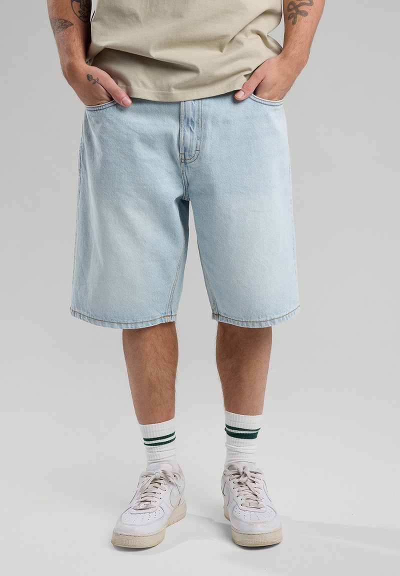 Vaaleansiniset farkkushortsit, joissa on väljä istuvuus, etutaskut, kevyesti kulunut pinta ja käännetty lahje. Yhdistetty valkoisiin tennareihin ja raidallisiin sukkiin.