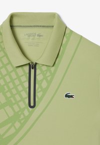 Polo de color verde claro con cuello de cremallera, con un patrón texturizado y un pequeño logo de cocodrilo bordado en la parte frontal.