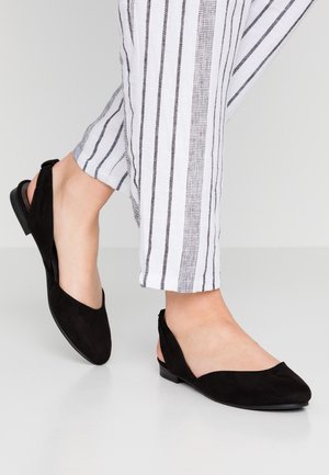 Pies con zapatos planos slingback de ante negro combinados con pantalones blancos con rayas negras verticales.
