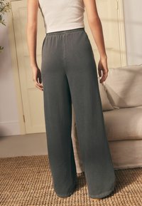 Persona che indossa pantaloni larghi grigi scuri e una camicetta bianca senza maniche, in piedi su un tappeto intrecciato vicino a un divano beige.