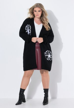 Cardigan nero lavorato a maglia con ricami floreali bianchi su entrambi i lati, con apertura frontale, maniche lunghe, indossato sopra una maglietta bianca e una gonna bordeaux.