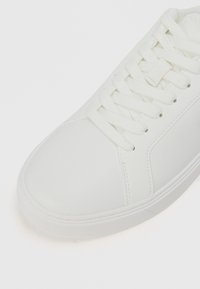 Białe syntetyczne sneakersy z płaskimi sznurówkami, okrągłym noskiem, teksturowanymi bocznymi panelami i gładkim wykończeniem. Smukły, minimalistyczny design.