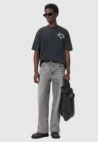 Černé volné tričko s bílou grafikou, šedé džíny s rovnými nohavicemi, robustní černé boty a černá taška s nastavitelným popruhem.