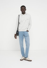 Γκρι βαμβακερή μπλούζα με κουκούλα και κορδόνια, ανοιχτό μπλε τζιν και μπεζ αθλητικά παπούτσια. Η μπλούζα με κουκούλα έχει ένα διακριτικό λογότυπο στο στήθος.