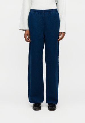 Osoba ubrana w ciemnoniebieskie szerokie spodnie z elastycznym pasem, czarne mokasyny i białą bluzkę z długim rękawem.