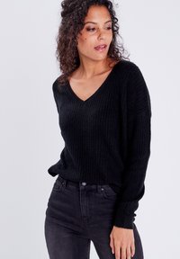 Schwarzer, gerippter Strickpullover mit V-Ausschnitt und langen Ärmeln, kombiniert mit dunklen Jeans. Der Stoff wirkt weich und hat eine strukturierte Oberfläche.