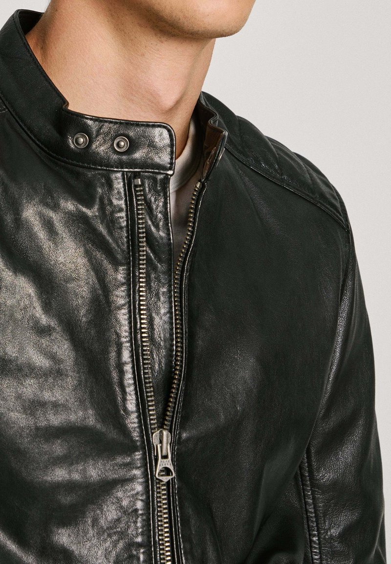 Chaqueta de cuero negra con cuello de botón y cierre de cremallera, con una textura suave y un detalle de hardware mínimo.