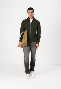 Grønn zip-up genser, grå jeans og hvite sneakers. Holder en beige tote bag. Minimalistisk design, glatt tekstur og avslappet passform.