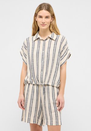 Frau trägt ein beige-blau gestreiftes Kurzarmhemd mit Knöpfen und passende Shorts mit Kordelzug und steht vor einem einfarbigen Hintergrund.