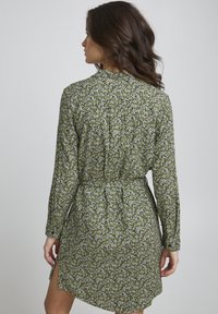 Abito floreale verde con maniche lunghe, motivo floreale che presenta fiori bianchi, legato in vita, lunghezza sotto il ginocchio e spacchi laterali.