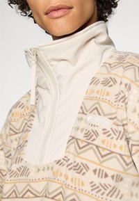 Krémový fleecový pulovr s geometrickým vzorem v hnědé, oranžové a zelené barvě. Má límec na poloviční zip a logo Columbia na hrudi.