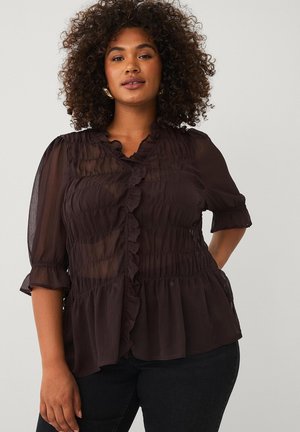 Vrouw met krullend haar, gekleed in een zwarte doorschijnende blouse met ruches en een zwarte broek, staand tegen een effen achtergrond.