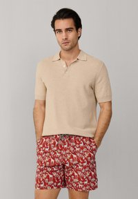 Homme portant un polo beige à manches courtes et un short de bain rouge avec des motifs blancs d'animaux et de plantes, debout les mains dans les poches.