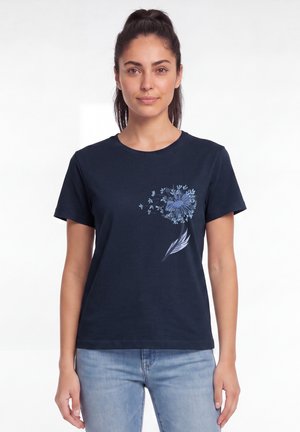 Femme aux cheveux foncés portant un t-shirt bleu marine avec un motif pissenlit bleu sur le côté gauche, associé à un jean bleu clair.