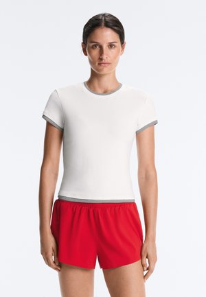 Femme aux cheveux foncés portant un t-shirt blanc à manches courtes avec un bord gris et un short de sport rouge, debout devant un fond uni.