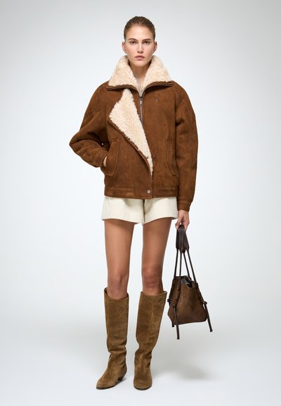 Bruine suede jas met een nepbont kraag, oversized pasvorm, voorkant met rits en drukknoopsluiting. Model draagt beige shorts en bruine knie-hoge laarzen.