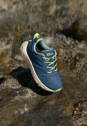 Chaussure de randonnée bleue avec un matériau synthétique texturé, des accents en mesh, des lacets vert fluo et une semelle blanche et robuste, posée sur une surface rocheuse.