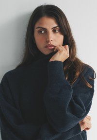 Žena s dlhými hnedými vlasmi v tmavomodrom svetri, ktorá drží golier blízko úst a zamyslene pozerá do blata na obyčajnej stene.