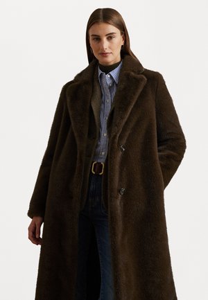 Manteau en fausse fourrure marron avec un grand col, design croisé et poches latérales, porté sur une chemise à rayures bleues et un jean en denim.
