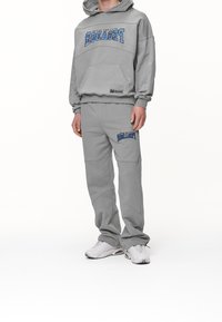 Grå hoodie med en framficka och blå grafisk text, parat med matchande joggingbyxor som har sidofickor och logodetaljer. Vita sneakers.