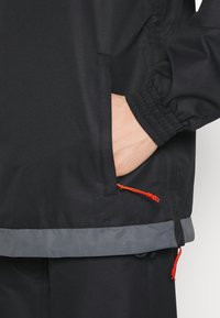 Musta takki, jossa on elastiset hihansuut ja harmaa alahelma, sivutasku oranssilla nyörillä ja sileä kangas.