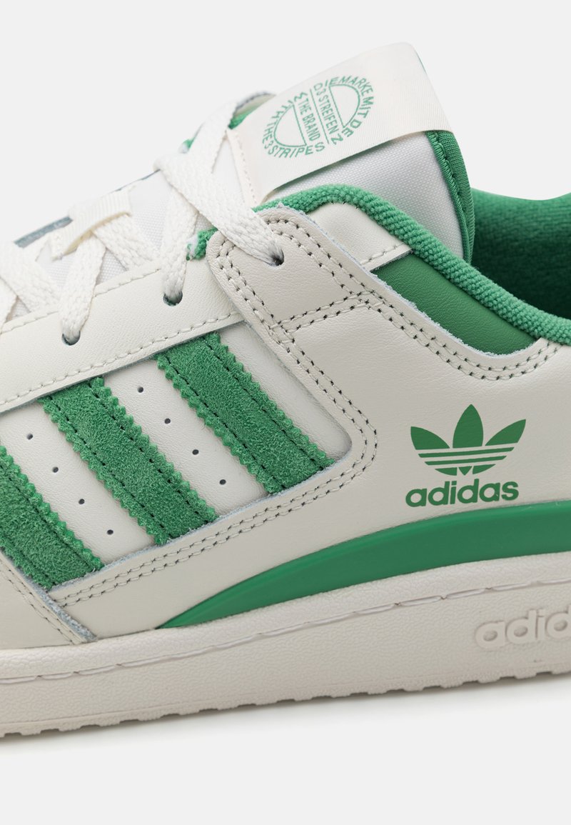 Zapatilla blanca de cuero con franjas y acentos de gamuza verde. Presenta cordones blancos, lengua texturizada y una suela de goma verde. Logo de Adidas presente.