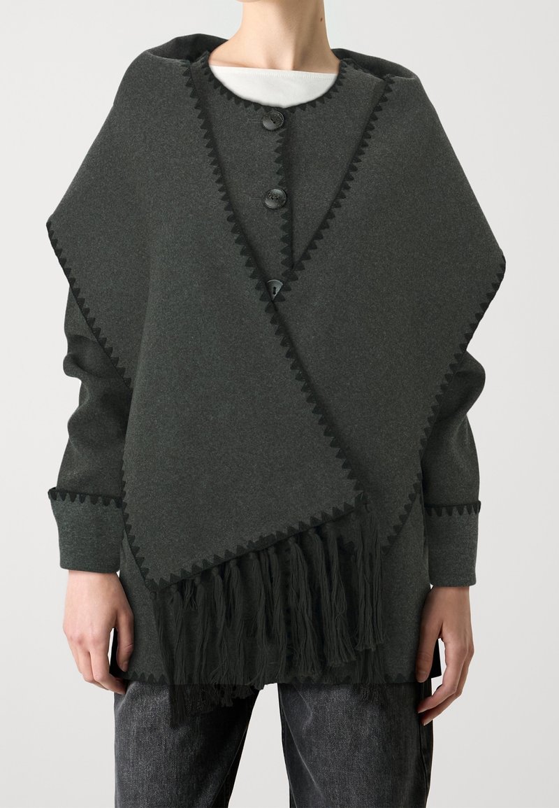 Poncho grigio scuro con orlo frangiato, chiusura a bottone e bordo a zigzag. Realizzato in morbido tessuto testurizzato con una vestibilità rilassata.